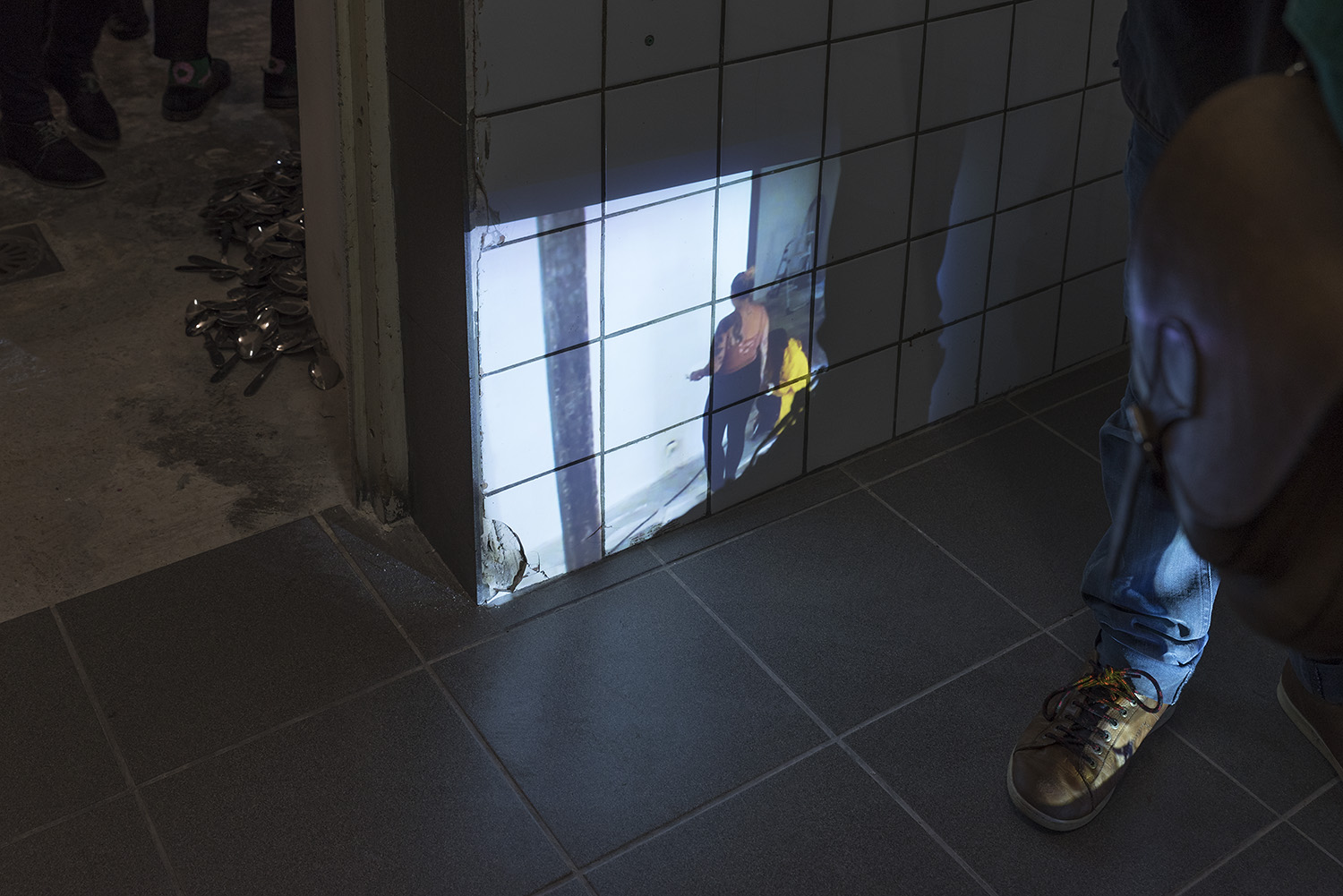 Dans un espace en carelage (mur et sol) on voit une image projeter sur le mur. Ce sont des gens en train de faire des travaux. Quelqu'un est posé devant la vidéo et son pieds cache une partie de la projection.